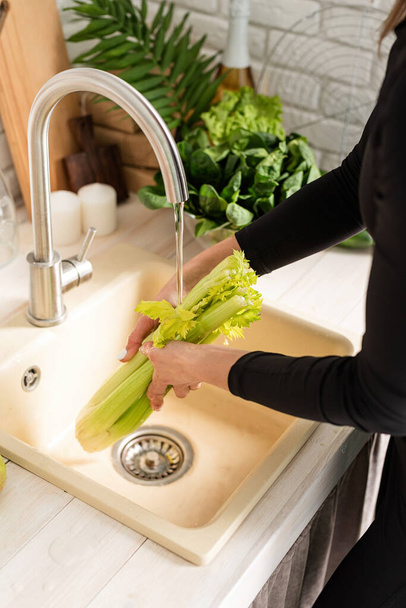 Preparing healthy foods. Young smiling woman in sport clothes washing celery in the kitchen - 写真・画像