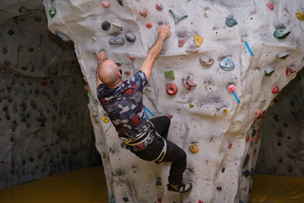 Active bald sporty man wearing protective face mask practicing rock climbing on artificial rock in a climbing wall. New normal in extreme sports. - Valokuva, kuva