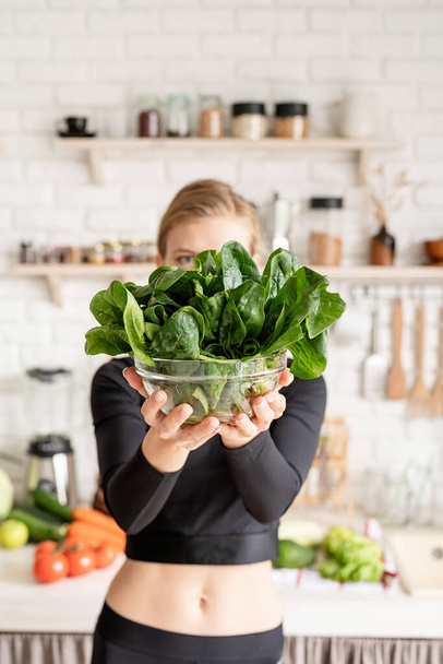 Healthy eating, dieting concept. Young blond smiling woman holding a bowl of fresh spinach in the kitchen - Foto, immagini