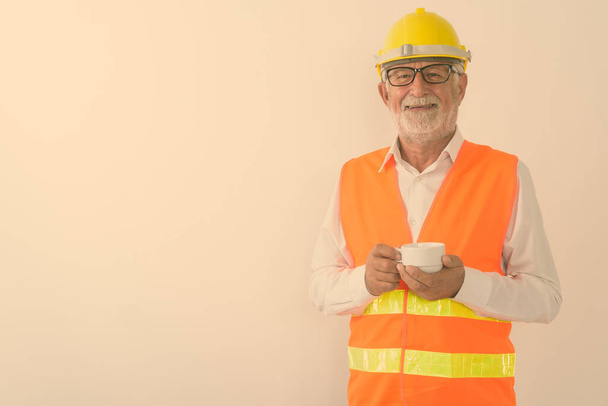 Studio strzał szczęśliwy starszy brodaty człowiek budowlany pracownik uśmiechając się i trzymając filiżankę kawy podczas noszenia okularów na białym tle - Zdjęcie, obraz