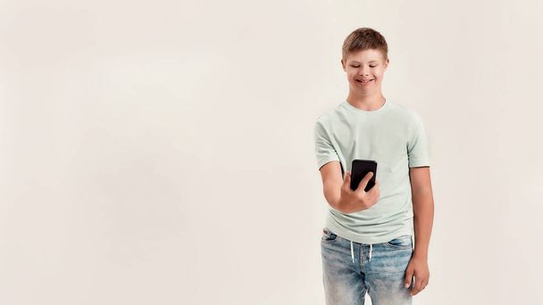 Happy disabled boy with Down syndrome holding smartphone, looking at the screen while standing isolated over white background - Fotografie, Obrázek