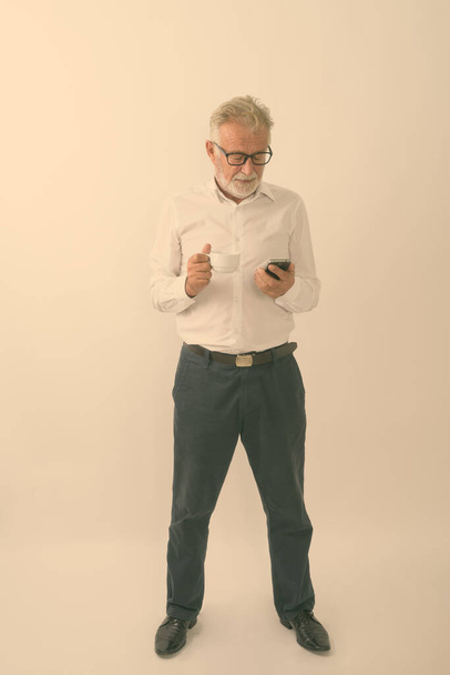 フルボディショットのハンサムな先輩ひげを生やした男立ちながら使用して携帯電話とホワイトの背景にコーヒーカップを保持 - 写真・画像