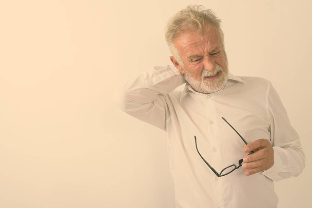 Studio záběr stresu senior vousatý muž s bolestí krku při držení brýlí proti bílému pozadí - Fotografie, Obrázek