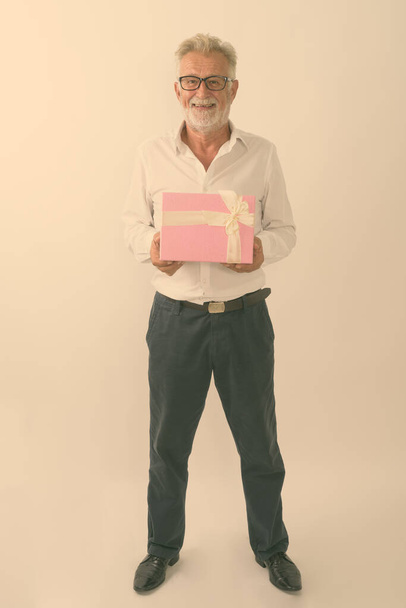 Full body shot of happy senior bearded man smiling and standing while holding gift box against white background - Фото, изображение