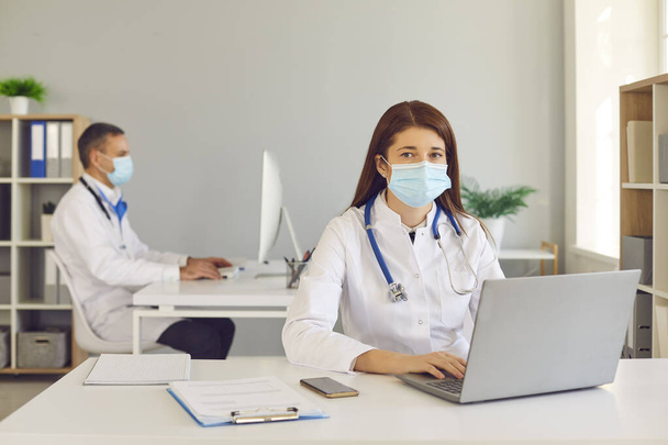 Young doctor in medical face mask working on laptop in hospital office together with colleague - Фото, зображення