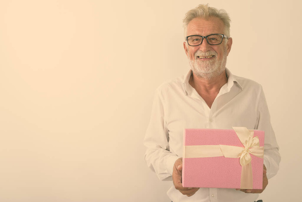 Studioaufnahme eines glücklichen älteren bärtigen Mannes, der lächelt, während er eine Geschenkbox mit Brille vor weißem Hintergrund hält - Foto, Bild