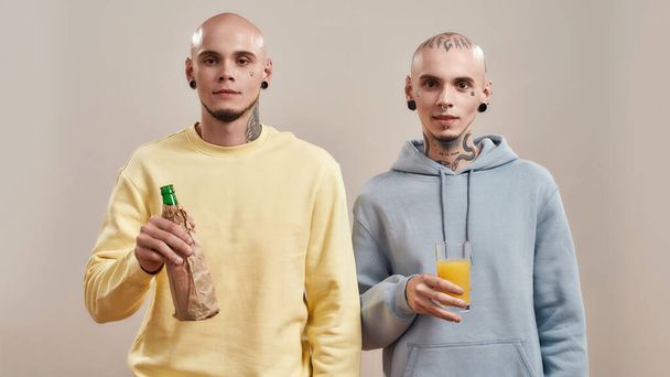 Healthy lifestyle. Young caucasian twin brothers in casual wear with tattoos and piercings holding beer bottle inside paper bag and glass of orange fresh - Fotó, kép