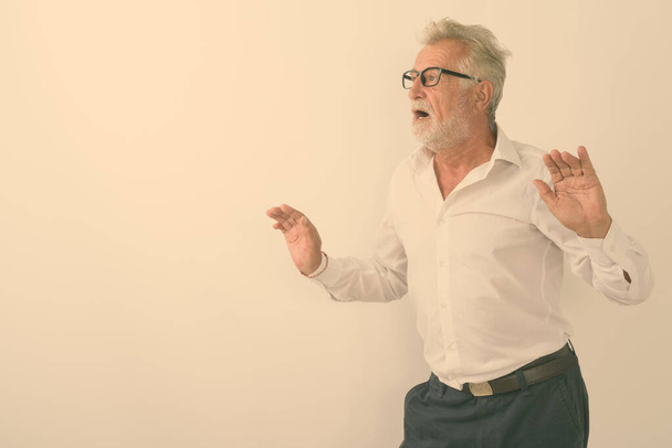 Studioaufnahme eines gutaussehenden älteren bärtigen Mannes, der überrascht mit erhobenen Armen aussieht, während er eine Brille vor weißem Hintergrund trägt - Foto, Bild