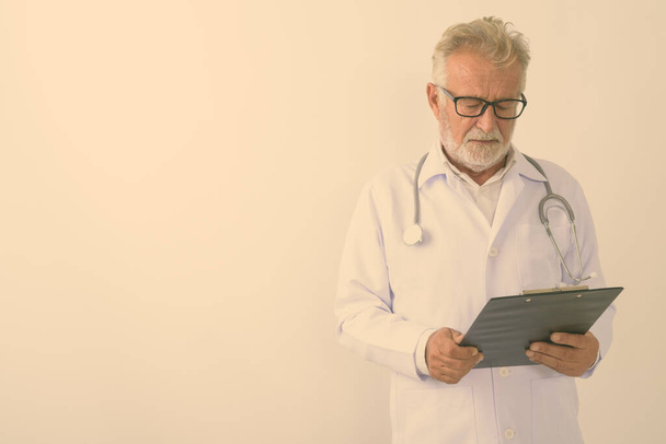 スタジオショットのハンサムなシニアひげそり男医師読書上のクリップボード上の白の背景 - 写真・画像