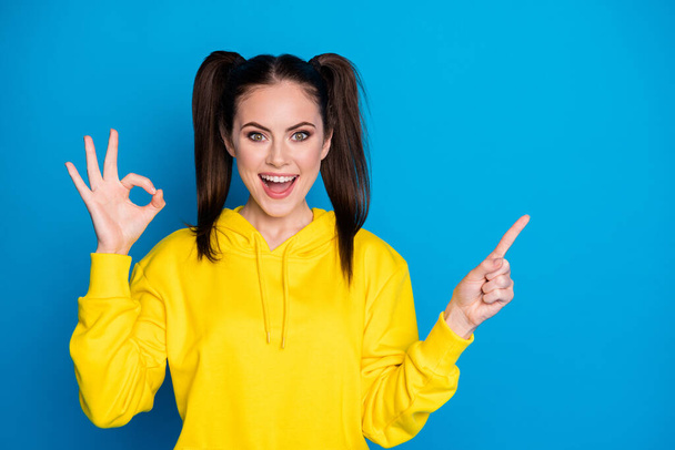 Photo of attractive lady two tails hairstyle direct fingers side empty space showing sale shopping banner okey symbol hand wear casual yellow hoodie pullover isolated blue color background - Fotó, kép