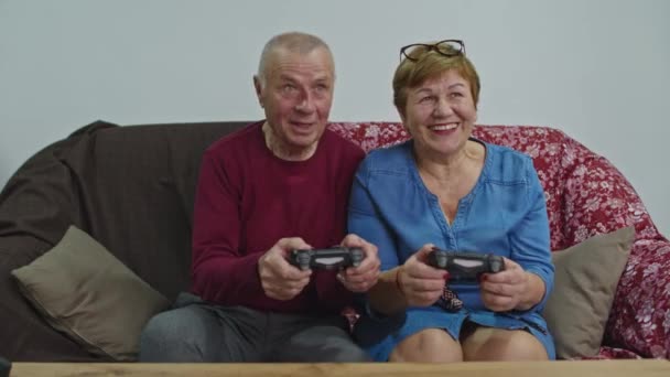 Ancianos pareja jugar con joysticks en casa en el sofá y sonreír felizmente. - Imágenes, Vídeo