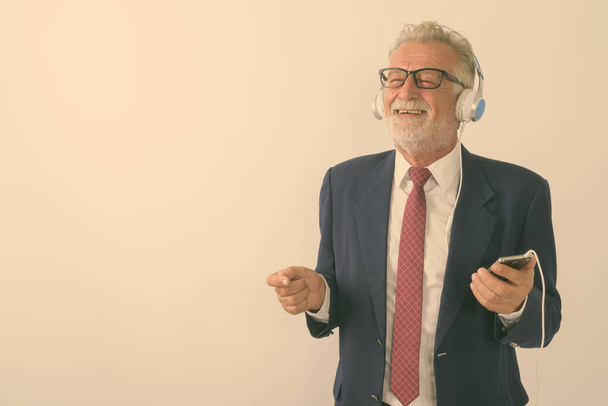 Studio shot of thoughtful happy senior bearded businessman smiling while holding mobile phone and listening to music against white background - Fotó, kép