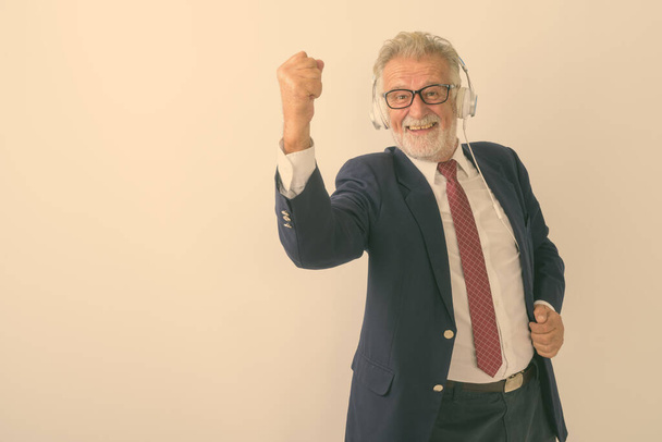 Studio shot of happy senior bearded businessman smiling while listening to music and looking motivated with eyeglasses against white background - Foto, immagini