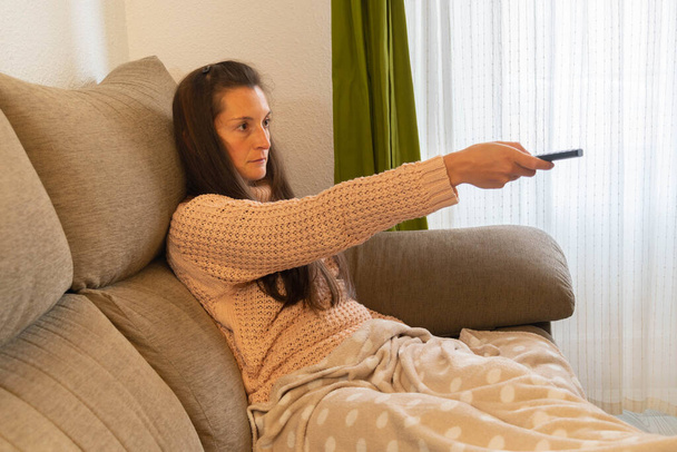 Femme sérieuse assise sur un canapé et recouverte d'une couverture change la chaîne avec la télécommande tout en regardant la télévision sérieusement - Photo, image