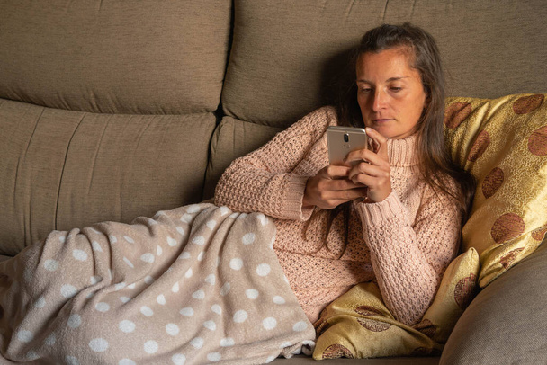 Woman lying on the sofa and covered with a blanket looks serious at her mobile phone, - Foto, Bild