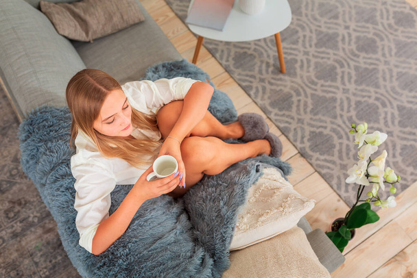Genç, güzel bir kız, kanepede, battaniyeye sarılmış, kahve ile oturuyor. - Fotoğraf, Görsel