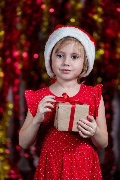 Adorable Little Girl In Santa Hat is Holding new year present with dreaming face looking at the camera. - Valokuva, kuva