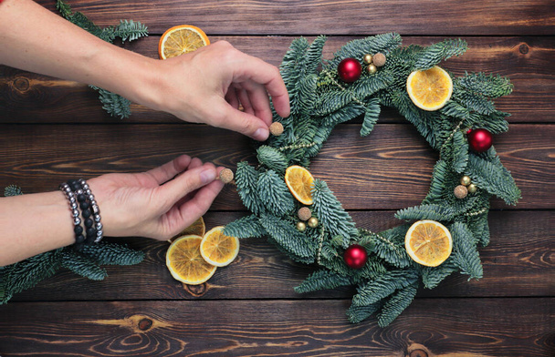 Naisten kädet koristavat kuusen oksista tehdyn joulun seppeleen, jossa on pieniä punaisia ja kultaisia palloja, kuivattuja oransseja, kiiltäviä helmiä puupöydällä. FLat lay. - Valokuva, kuva