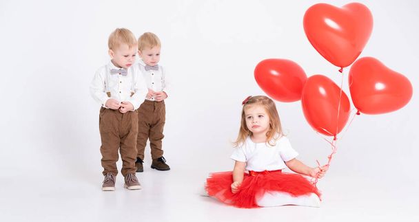zwei kleine Jungen in Anzügen schauen auf ein kleines Mädchen mit Herzballons auf weißem Hintergrund - Foto, Bild