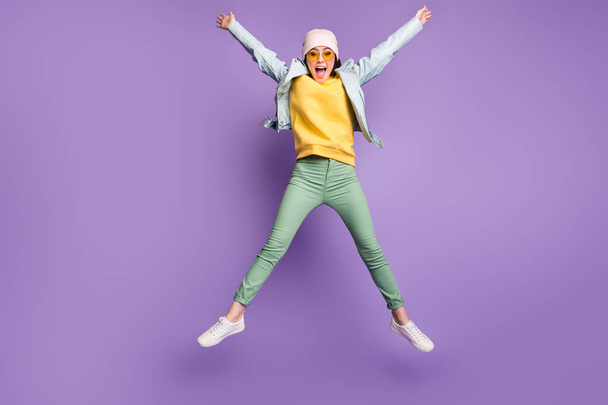 Full body photo of funky pretty lady stylish clothes jump high rejoicing spread hands star shape wear sun specs casual hat modern jacket green pants footwear isolated purple color background - Foto, Bild