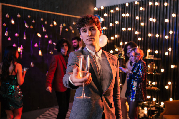 Attraente e divertente ragazzo con i capelli ricci che tiene un bicchiere di champagne davanti alla telecamera alla grande festa si gode il momento - Foto, immagini