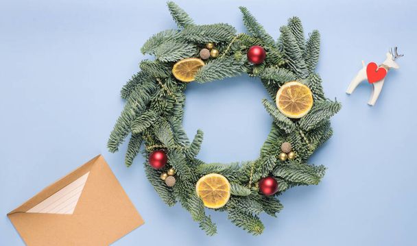 小さな赤と金のボール、乾燥オレンジのスライス、青い表面に光沢のあるビーズで作られたモミの枝で作られたクリスマスの花輪.  - 写真・画像