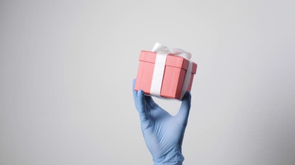 primer plano en la mano en guante protector azul que sostiene la caja de regalo roja con cinta blanca - Imágenes, Vídeo