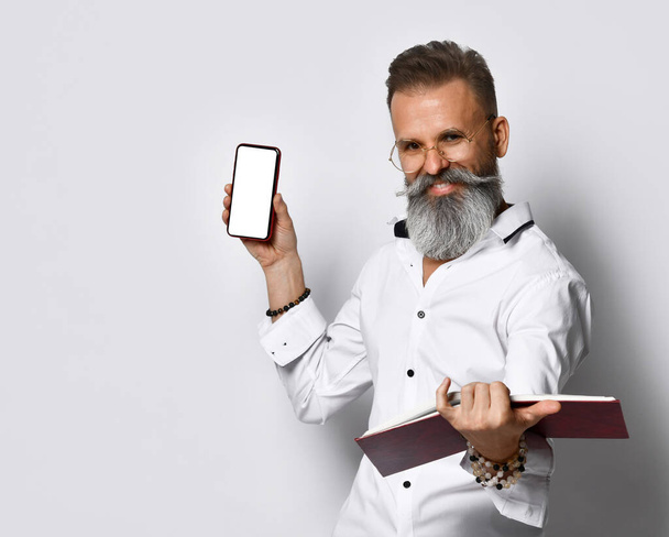 Smiling hipster man in eyewear with notebook showing smartphone - Zdjęcie, obraz