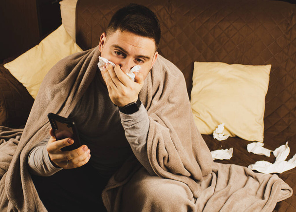 Kranker junger Mann sitzt in seinem Wohnzimmer und bläst Rotz und hat ein Thermometer im Mund, um sein Fieber zu messen - Foto, Bild