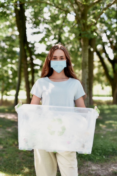 Young female eco activist wearing protective face mask holding recycle bin and looking at camera while collecting plastic waster in the forest during covid 19 pandemic - Fotografie, Obrázek