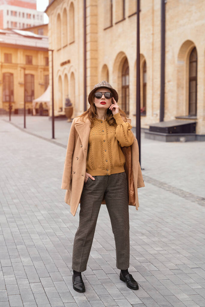 Full height self confident girl in an autumn beige coat and sunglasses, plaid panama hat standing posing on the street with coat on her shoulders. Retro toned photo - Fotoğraf, Görsel