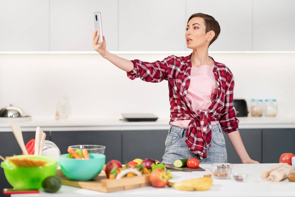 Pretty woman taking selfie or making a video call using her smartphone holding it in outstretched arm while cooking fresh salad wearing a plaid shirt with a bob hair style - Φωτογραφία, εικόνα