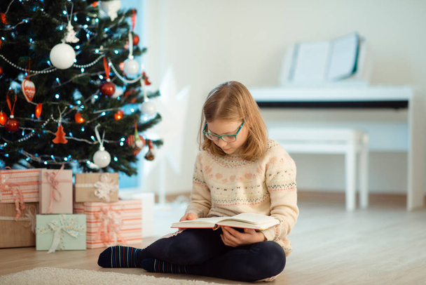 Portrait of pretty little child girl reading book sitting near decorated christmas tree - Foto, imagen