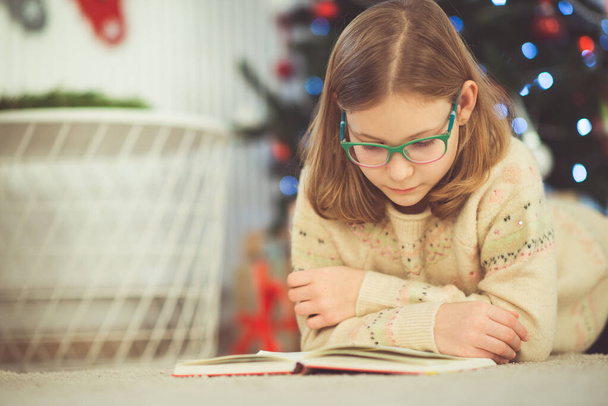 Portrait of pretty little child girl reading book sitting near decorated christmas tree - Valokuva, kuva