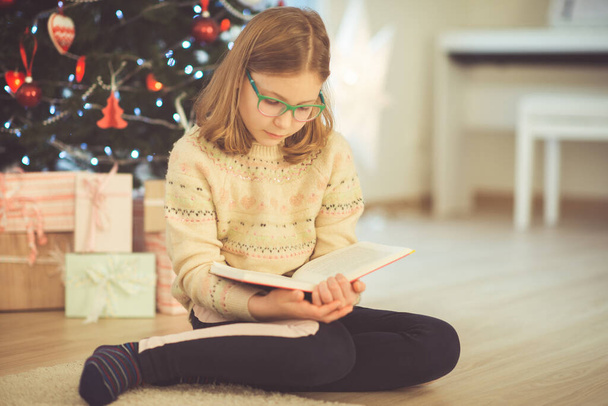 Portrait of pretty little child girl reading book sitting near decorated christmas tree - Photo, Image