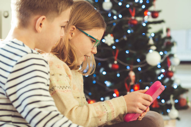 Happy teen siblings children or friends playing games with tablet on sofa near christmas tree at home - Φωτογραφία, εικόνα