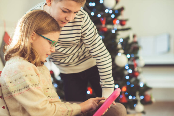 Happy teen siblings children or friends playing games with tablet on sofa near christmas tree at home - Foto, Imagen