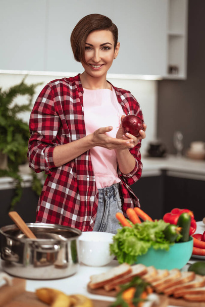 Dona de casa prepara o jantar para a família escolher um legumes segurando uma cebola vermelha em uma mão de pé na cozinha. Comida saudável em casa. Conceito de alimentação saudável - Foto, Imagem