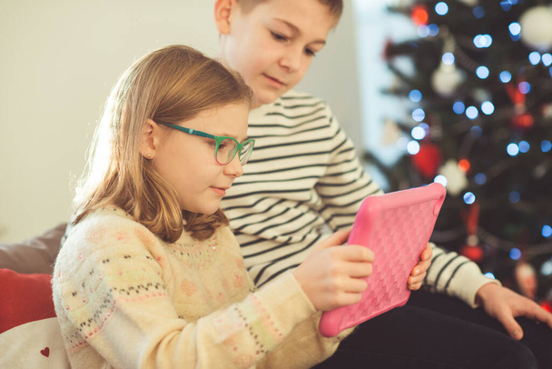 Feliz adolescente hermanos niños o amigos jugando juegos con la tableta en el sofá cerca del árbol de Navidad en casa - Foto, Imagen