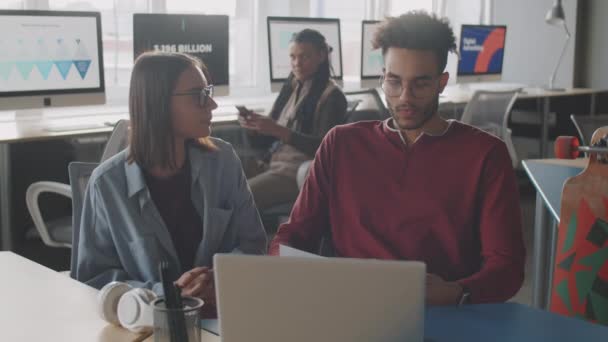 Joven hombre de raza mixta discutiendo documento de negocios con colega mujer caucásica mientras coopera durante la jornada laboral en la oficina - Metraje, vídeo