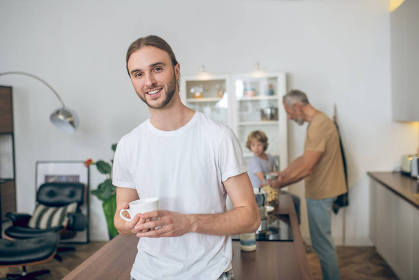 Young man in white tshirt standing in the kitchen and smiling - Фото, изображение