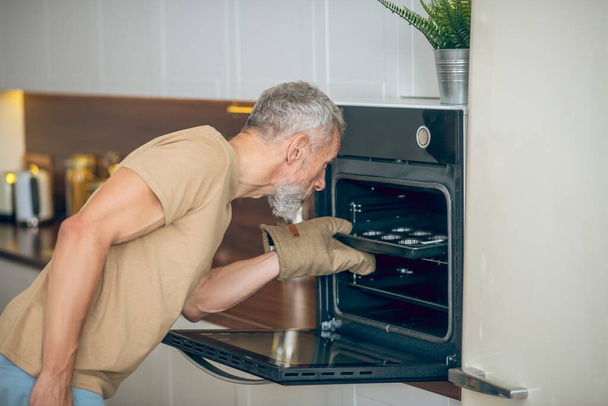 Mature man in beige tshirt taking something out the oven - Foto, Imagen