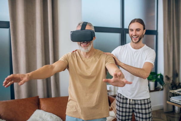 Mature man in vr glasses walking in the room, his partner supporting him - Foto, Imagem