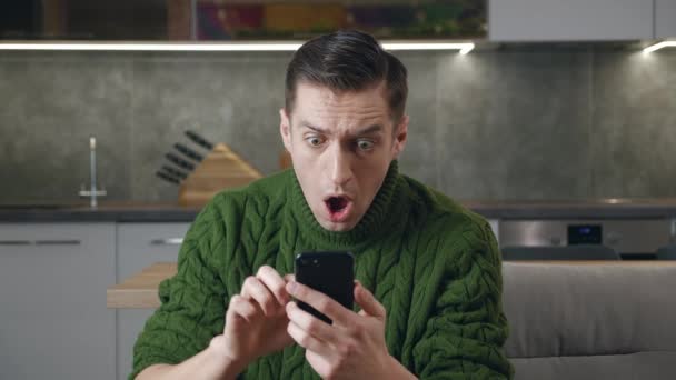 Šokovaný muž reagující na hroznou zprávu na smartphonu si zakryje ústa hrůzou, když sedí doma na pohovce v obývacím pokoji. Nepříjemně překvapený a vystrašený muž čte špatné zprávy na mobilním telefonu - Záběry, video