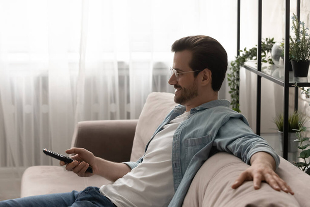 Relaxed young man watching tv choosing channel using remote control - Foto, Imagen