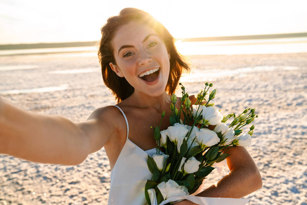 Cheery optimistic happy young beautiful woman at the beach taking a selfie in a beautiful sunny morning, holding bouquet - Фото, изображение