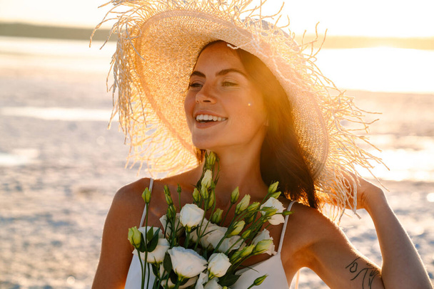Photo of a cheery optimistic happy young beautiful woman at the beach walking in a beautiful sunny morning, holding bouquet - Фото, изображение