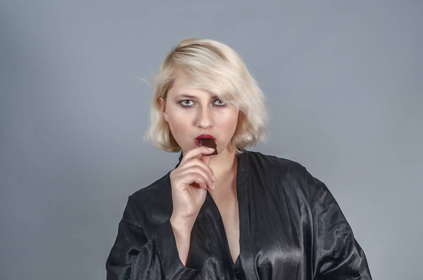 Attraktive junge blonde Frau, die vor grauem Hintergrund Schokolade isst. - Foto, Bild