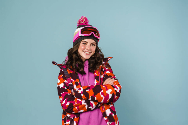 Happy young woman wearing snowboarding gear standing isolated over blue background - Valokuva, kuva