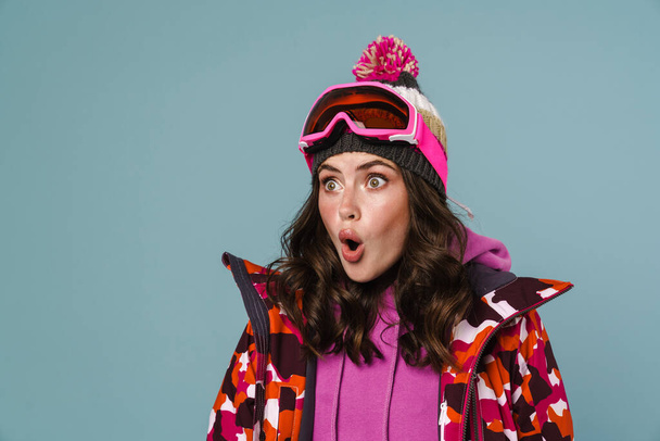 Shocked young woman wearing snowboard jacket isolated over blue background - 写真・画像